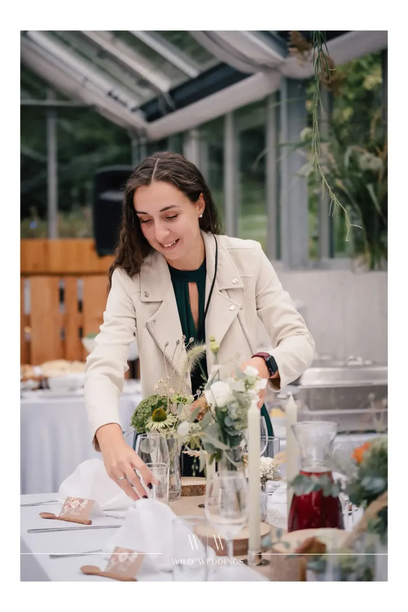 Floristin arrangiert Blumen auf festlich gedeckter Tafel im hellen Glashaus