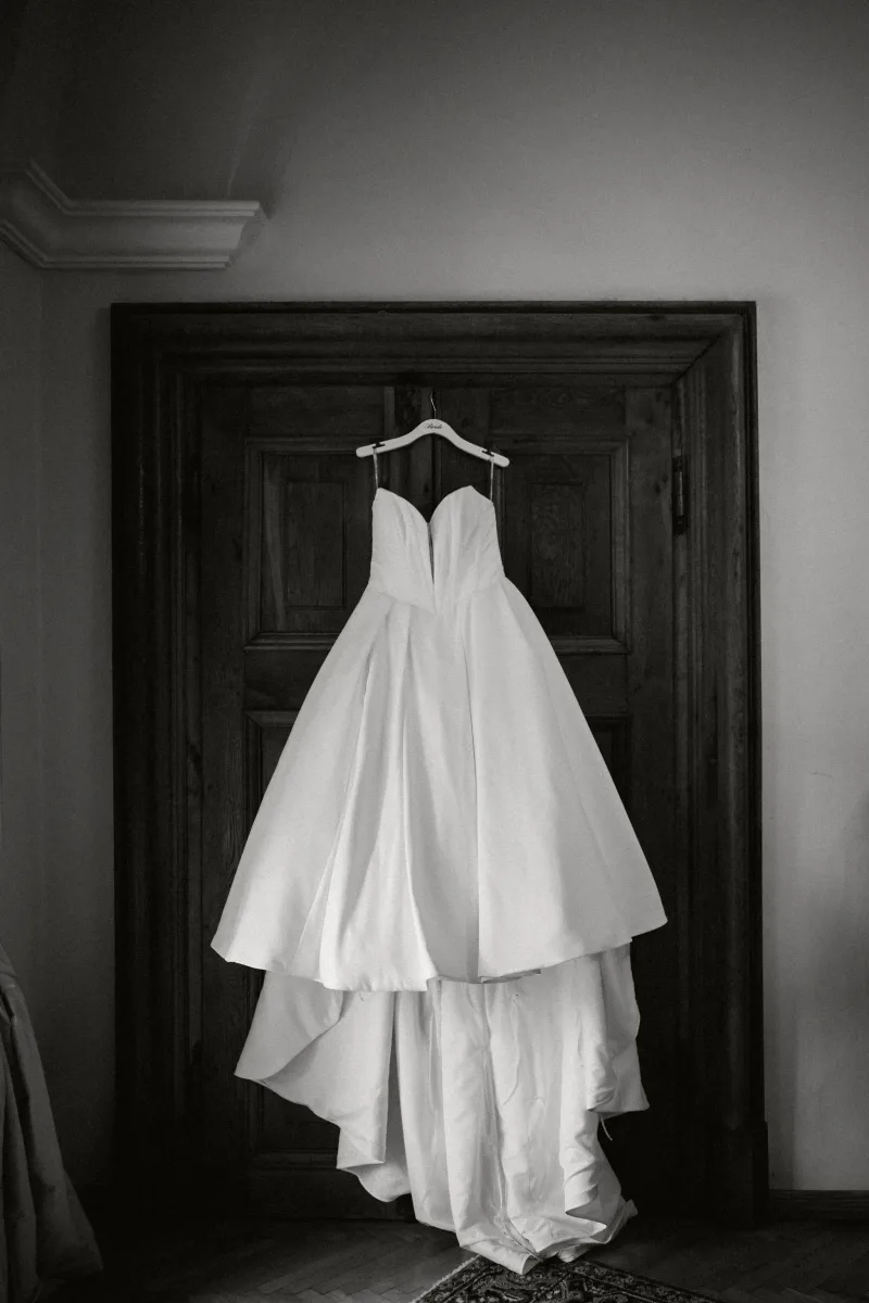 Weißes Brautkleid mit Schleppe hängt an einer dunklen Holztür in Schwarzweiß
