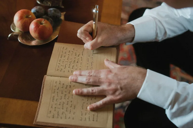 Hände schreiben in ein offenes Notizbuch mit goldenem Stift auf dunklem Holztisch