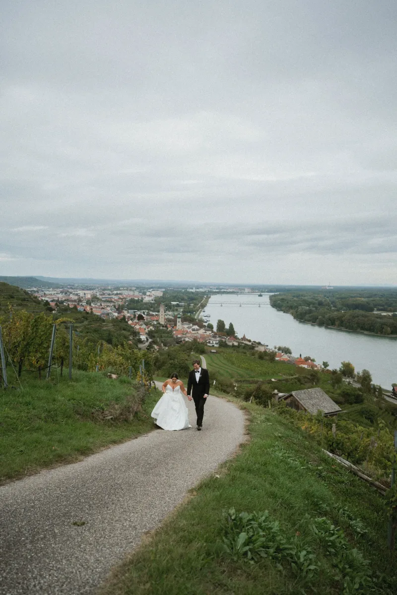 Brautpaar spaziert Hand in Hand über Kiesweg mit Stadt- und Flussblick im Hintergrund